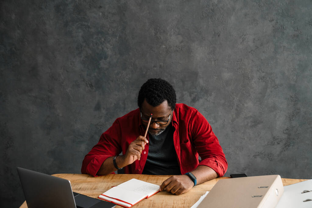 Черный человек в очках записывает заметки во время работы с ноутбуком в помещении - Фото, изображение