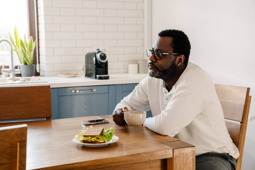 Black man wearing eyeglasses drinking coffee while having dinner at home - Foto, afbeelding
