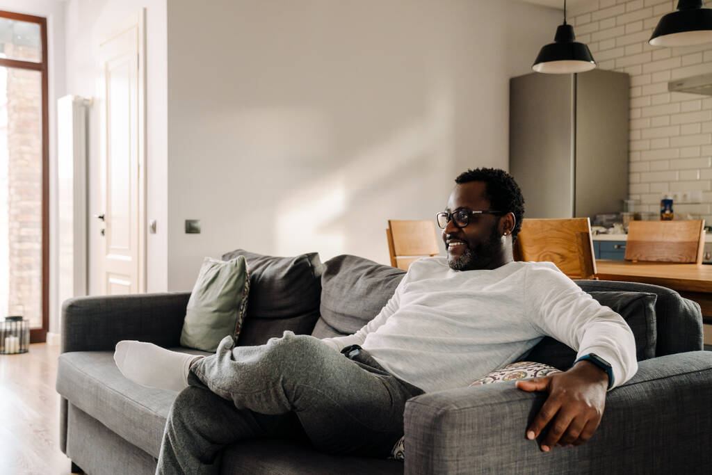 Чёрный мужчина в очках смотрит телевизор, отдыхая дома на диване. - Фото, изображение