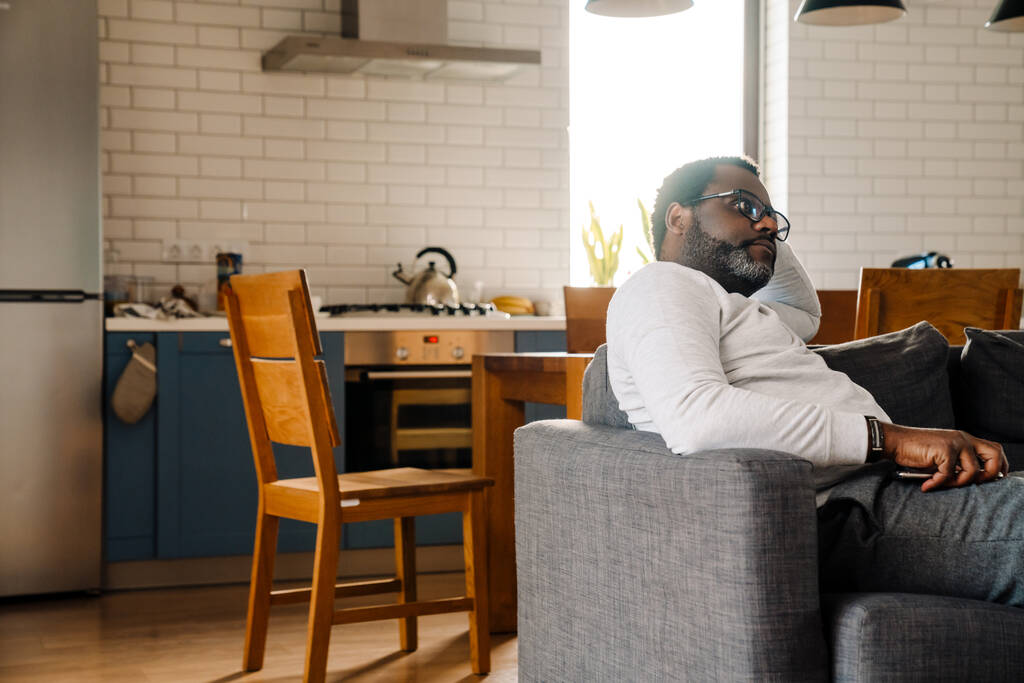Чорний чоловік в окулярах дивиться телевізор, відпочиваючи на дивані вдома
 - Фото, зображення
