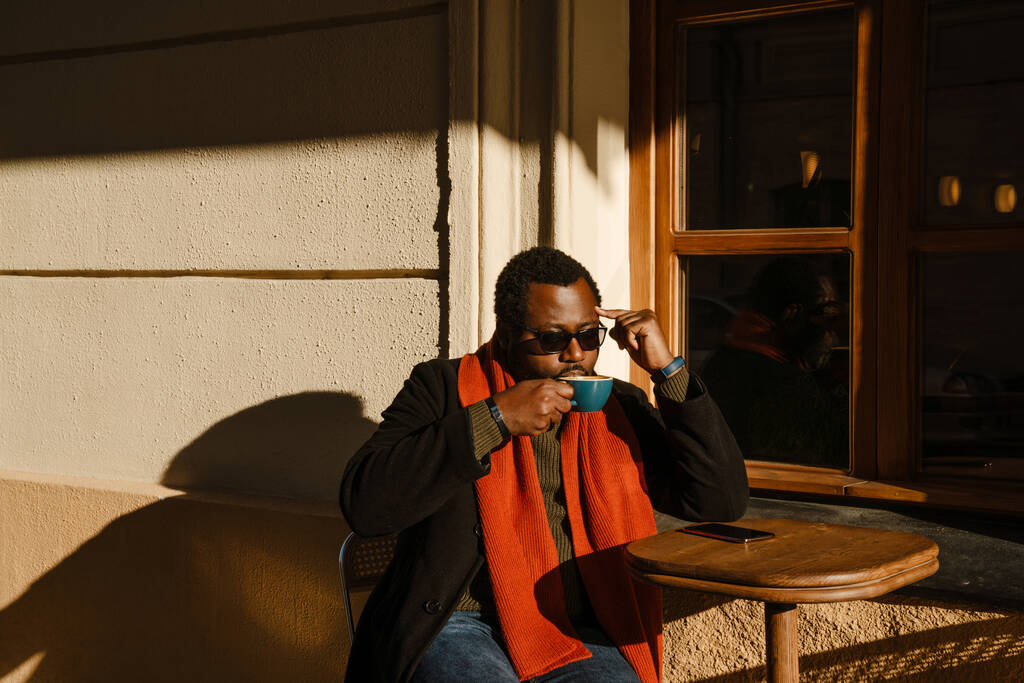 Black bearded man wearing sunglasses drinking coffee while sitting in street cafe outdoors - Foto, Imagem
