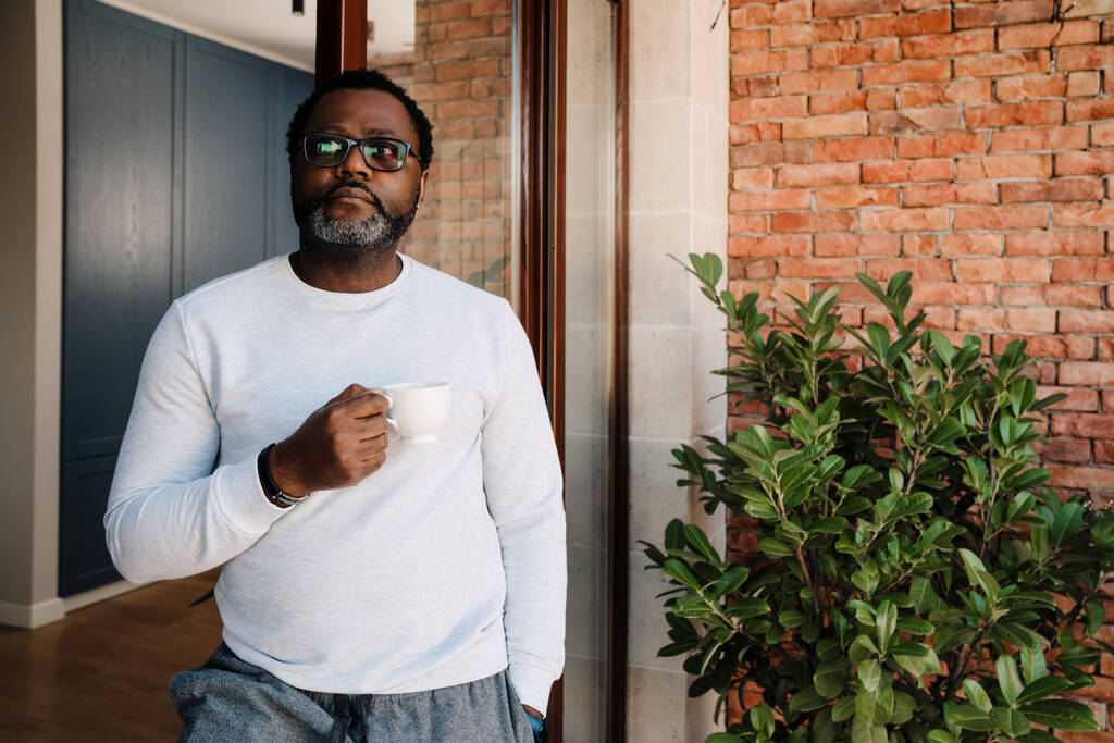 Black bearded man drinking coffee while standing in doorway at home - Foto, afbeelding