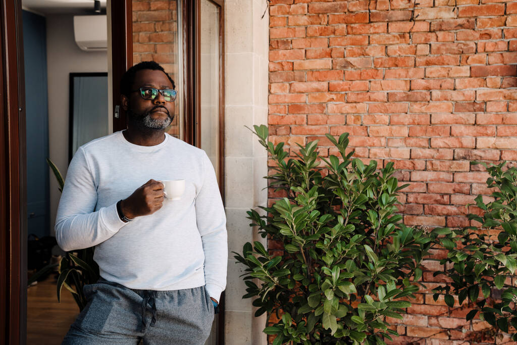 Black bearded man drinking coffee while standing in doorway at home - Foto, imagen