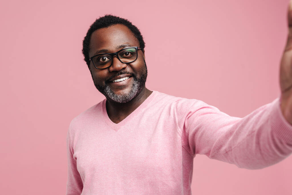 Black bearded man in eyeglasses smiling while taking selfie photo isolated over pink background - Foto, Bild