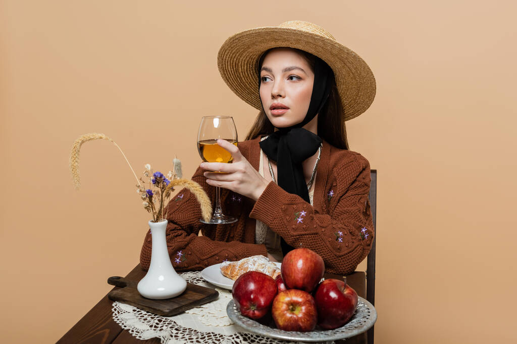 Stylish woman in sun hat holding wine near food on table isolated on beige  - Foto, Imagem