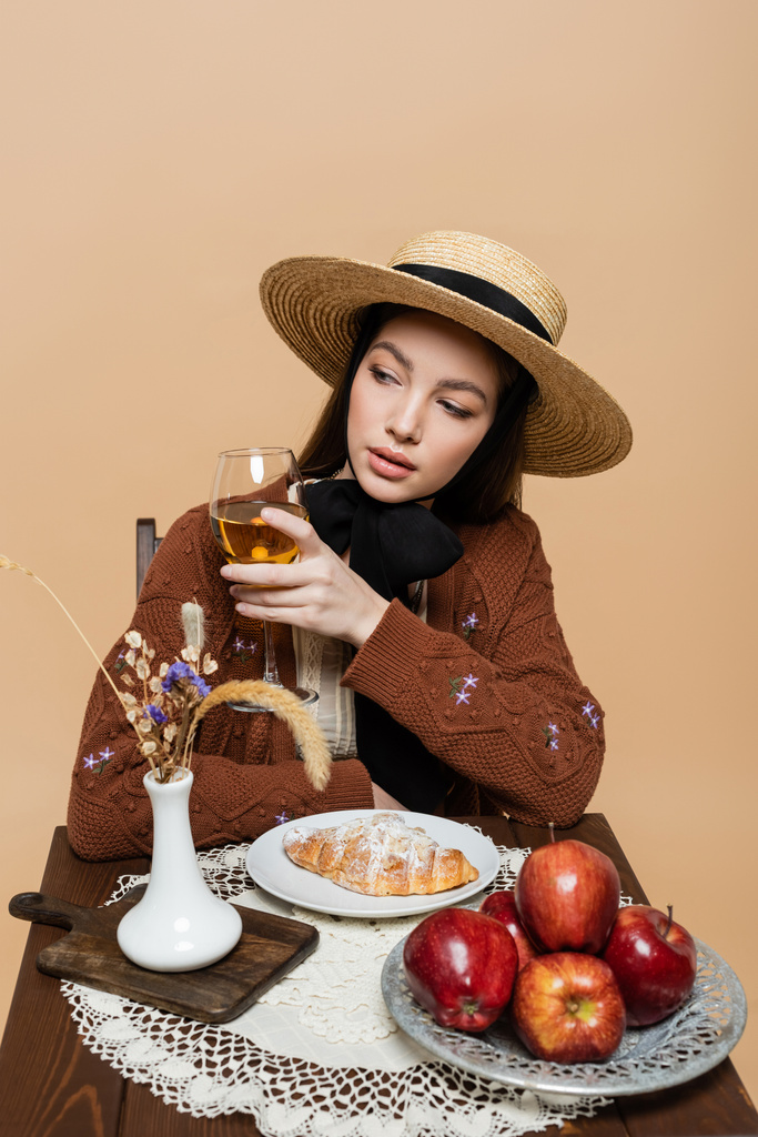 Trendy woman holding wine near apples and croissant on table isolated on beige  - Foto, immagini