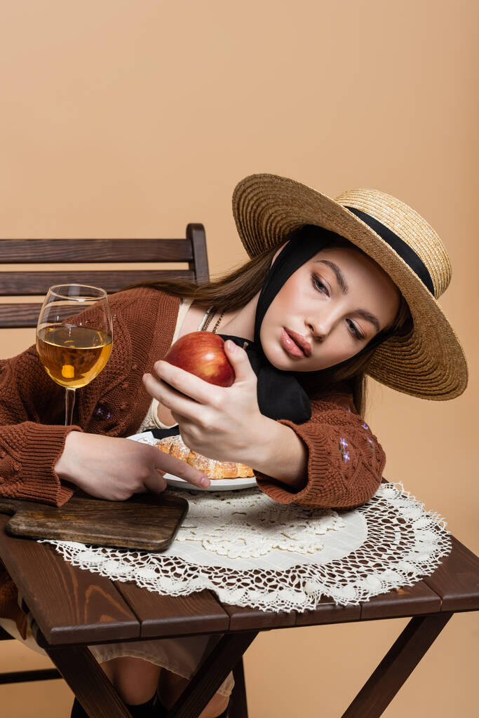 Stylish woman in sun hat holding apple near wine and croissant isolated on beige  - 写真・画像