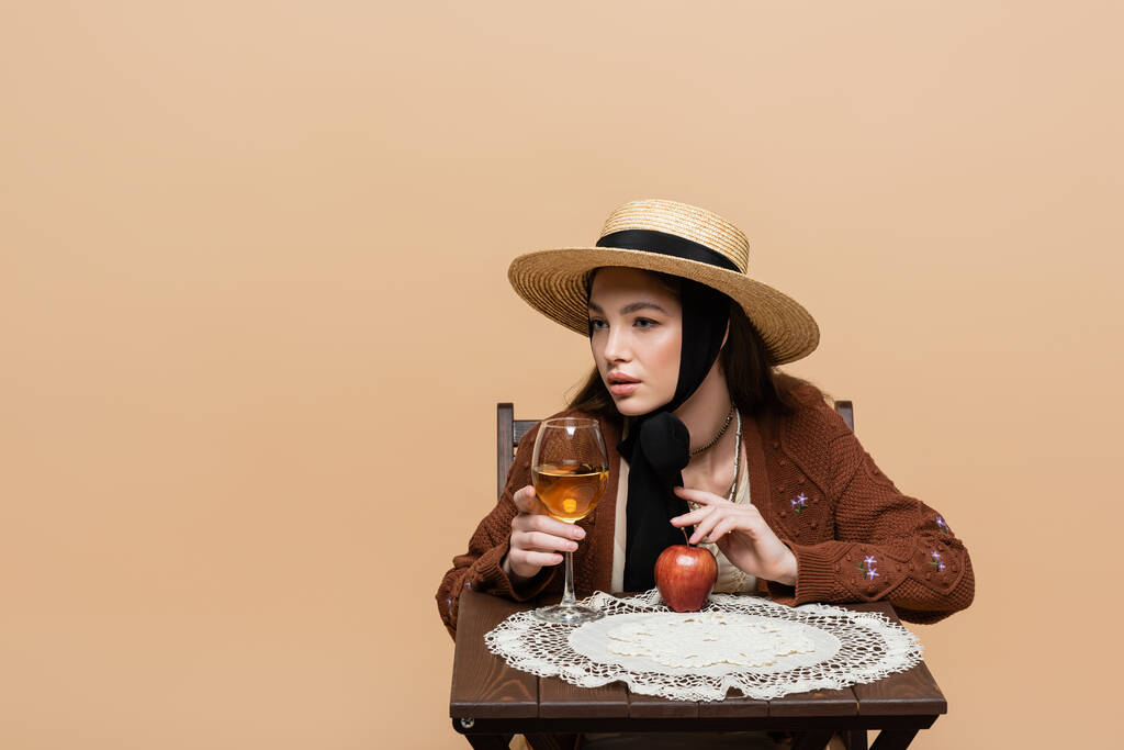 Молодая женщина в солнечной шляпе касается бокала вина и яблока на столе изолированы на бежевый  - Фото, изображение