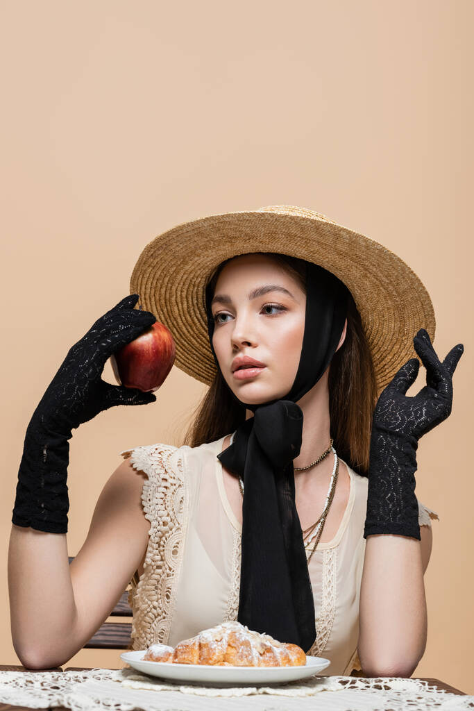 Модная женщина в соломенной шляпе и перчатках держит яблоко возле круассана изолированы на бежевый  - Фото, изображение