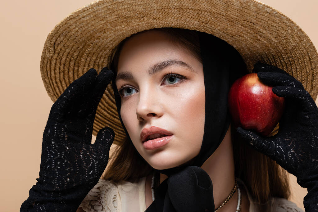 Портрет красивой женщины в перчатках и соломенной шляпе, держащей яблоко, изолированное на бежевом  - Фото, изображение