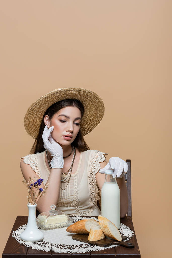 Модная модель в солнцезащитной шляпе трогательная бутылка молока рядом с маслом и хлеб изолирован на бежевый  - Фото, изображение