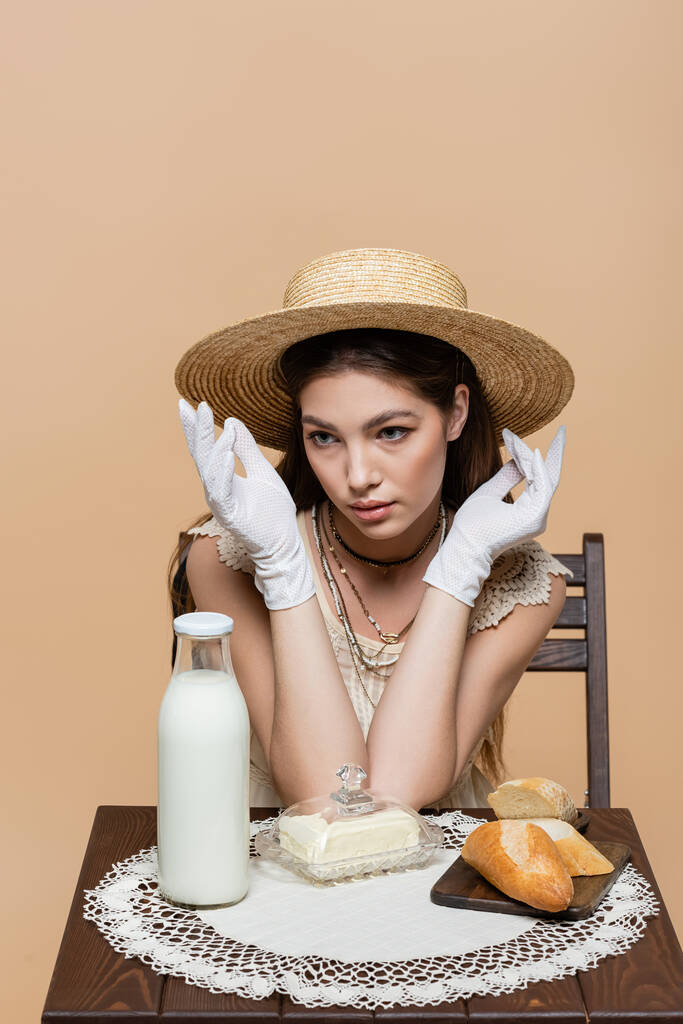 Молодая женщина в перчатках и шляпе, сидя возле бутылки молока и хлеба изолированы на бежевый  - Фото, изображение