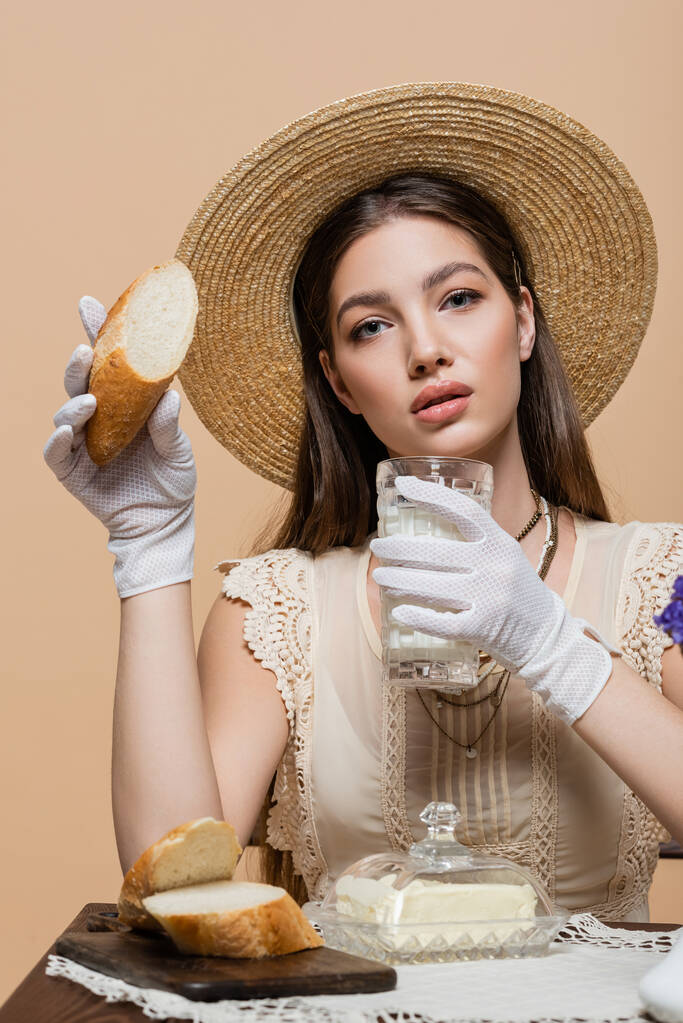 Πορτρέτο της κομψής γυναίκας στο καπέλο ήλιο κρατώντας το γάλα και το ψωμί κοντά στο βούτυρο που απομονώνονται σε μπεζ  - Φωτογραφία, εικόνα