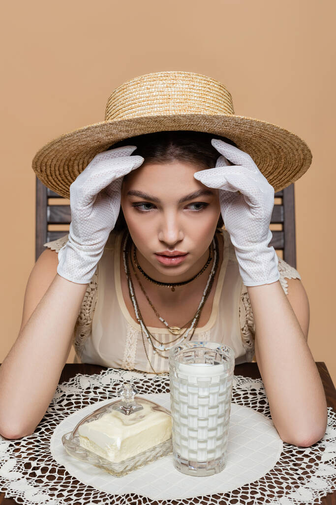 Pretty woman in sun hat looking away near milk and butter on table isolated on beige  - Photo, image