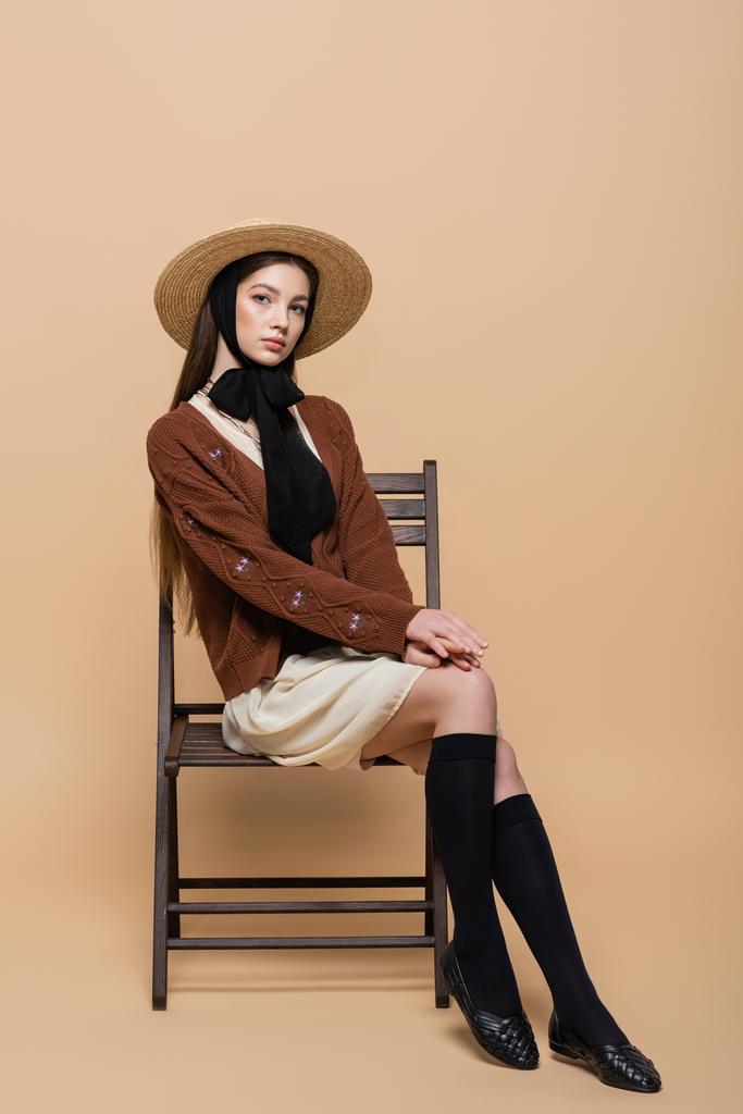 Молодая модель в соломенной шляпе сидит на стуле на бежевом фоне  - Фото, изображение