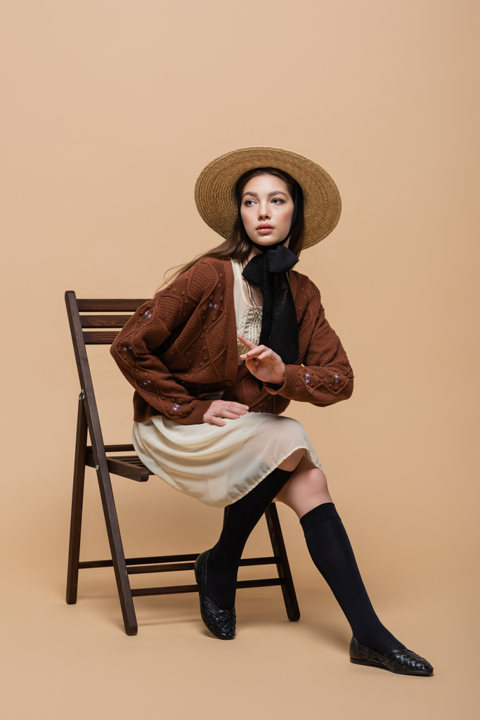 Pretty and stylish woman in sun hat pointing with finger while sitting on chair on beige background  - Fotó, kép