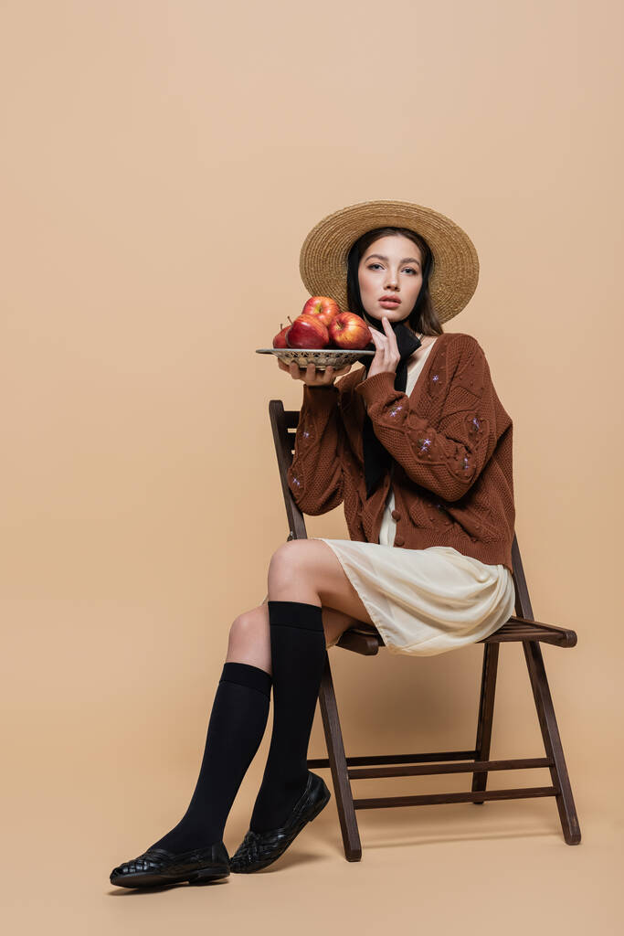 Trendy woman in sun hat holding fresh apples while sitting on chair on beige background  - Photo, image