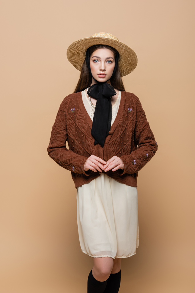 Pretty woman in knitted cardigan and straw hat looking at camera isolated on beige  - Photo, image