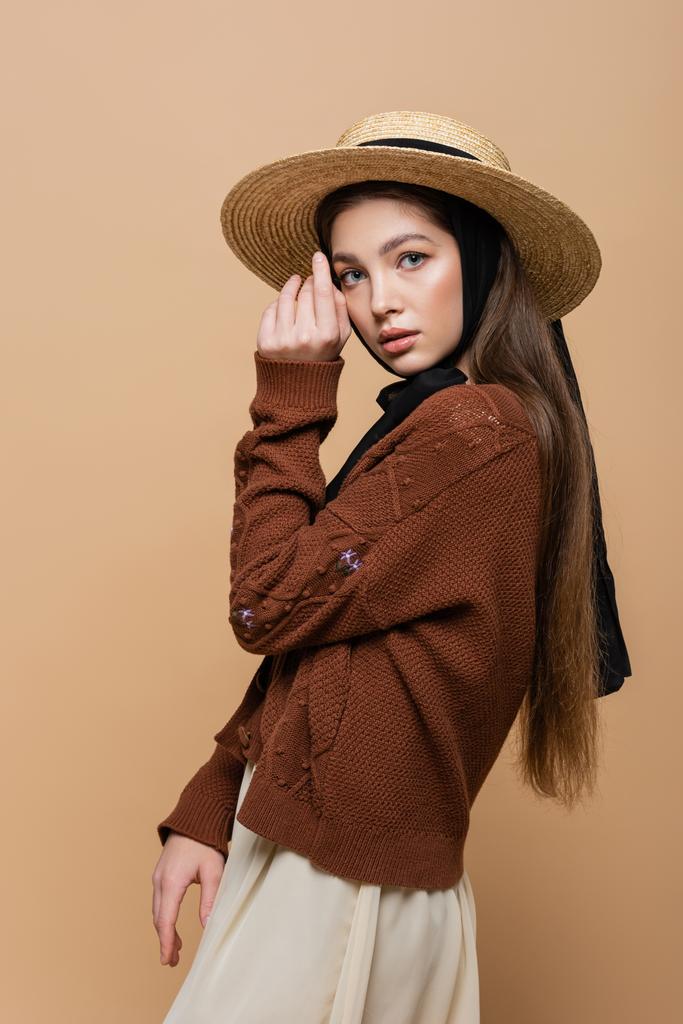Brunette woman in straw hat and knitted cardigan looking at camera isolated on beige  - Fotografie, Obrázek