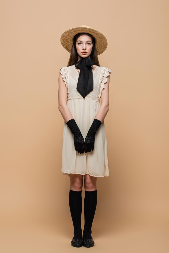 Πλήρες μήκος της γυναίκας σε ρετρό ρούχα και καπέλο ήλιο στέκεται σε μπεζ φόντο  - Φωτογραφία, εικόνα