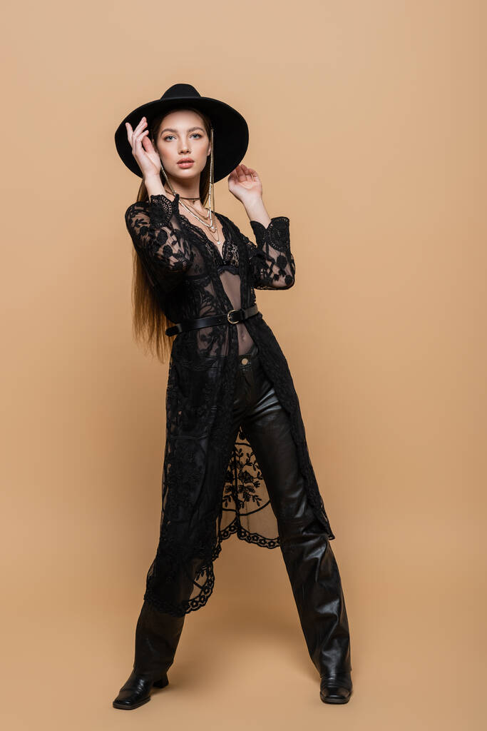 Stylish model in black hat and guipure robe looking at camera on beige background  - Foto, imagen