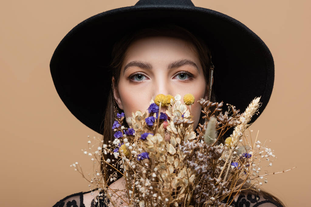Young woman in fedora hat looking at camera near dry flowers isolated on beige  - Photo, image