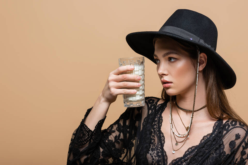 Trendy woman in fedora hat and guipure robe looking at glass of milk isolated on beige  - Foto, Bild