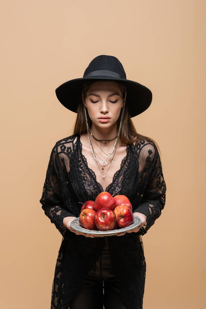 Trendy young woman in fedora hat holding fresh apples isolated on beige  - Photo, image