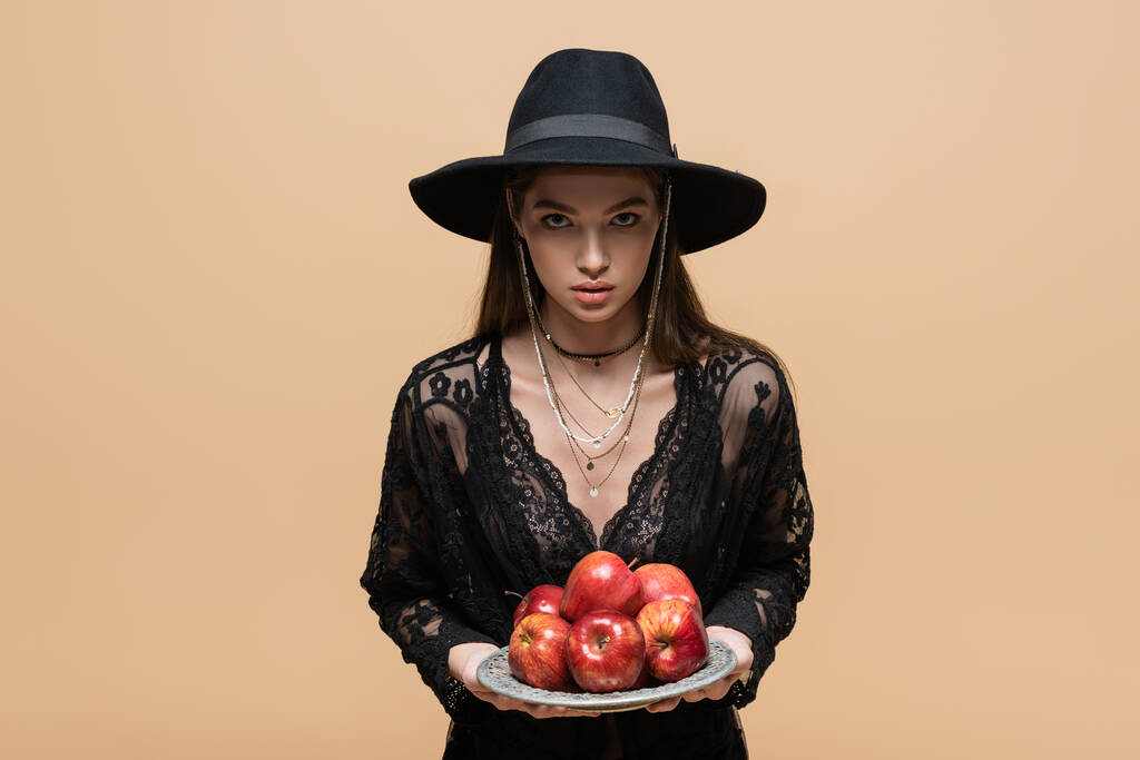 Stylish woman in fedora hat holding organic apples and looking at camera isolated on beige  - Fotó, kép