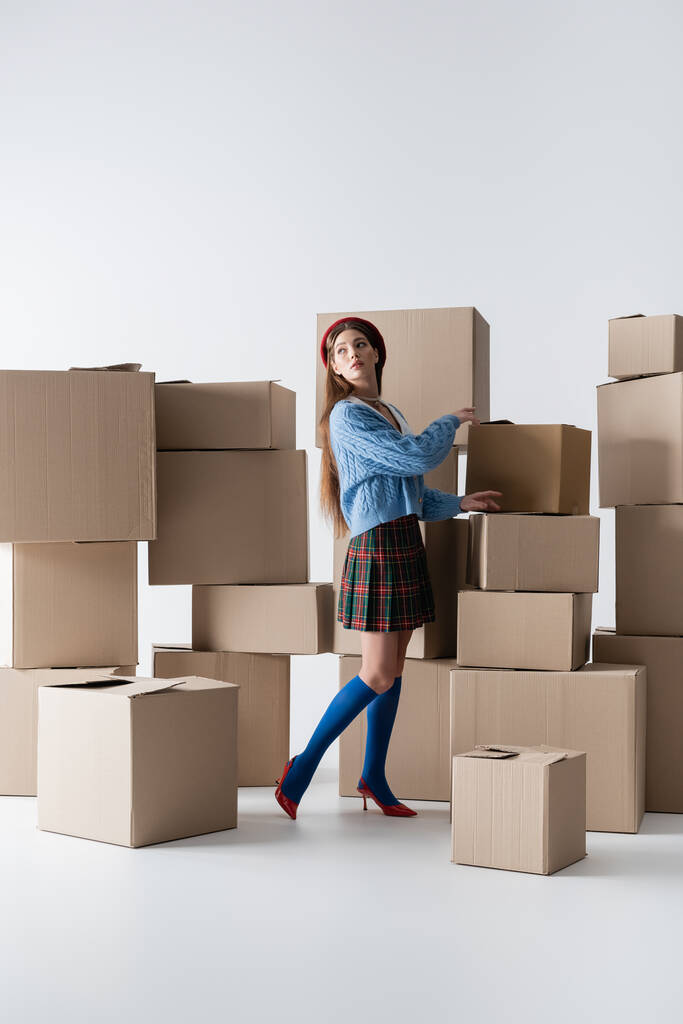Trendy woman in plaid skirt and cardigan looking away near carton boxes on white background  - Foto, Bild