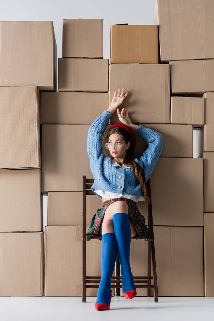 Trendy young woman n eyeglasses and beret sitting on chair near cardboard boxes on white background  - Fotó, kép