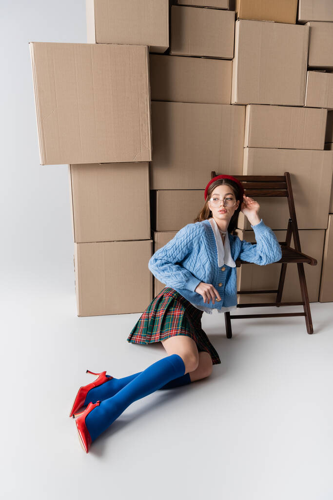 Stylish woman in heels and plaid skirt sitting near chair and cardboard boxes on white background  - Foto, immagini