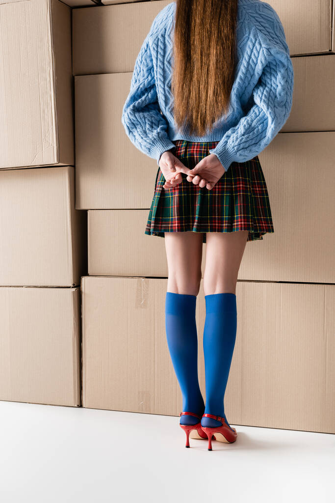 Cropped view of brunette woman in plaid skirt and heels standing near cardboard boxes on white background  - Fotografie, Obrázek