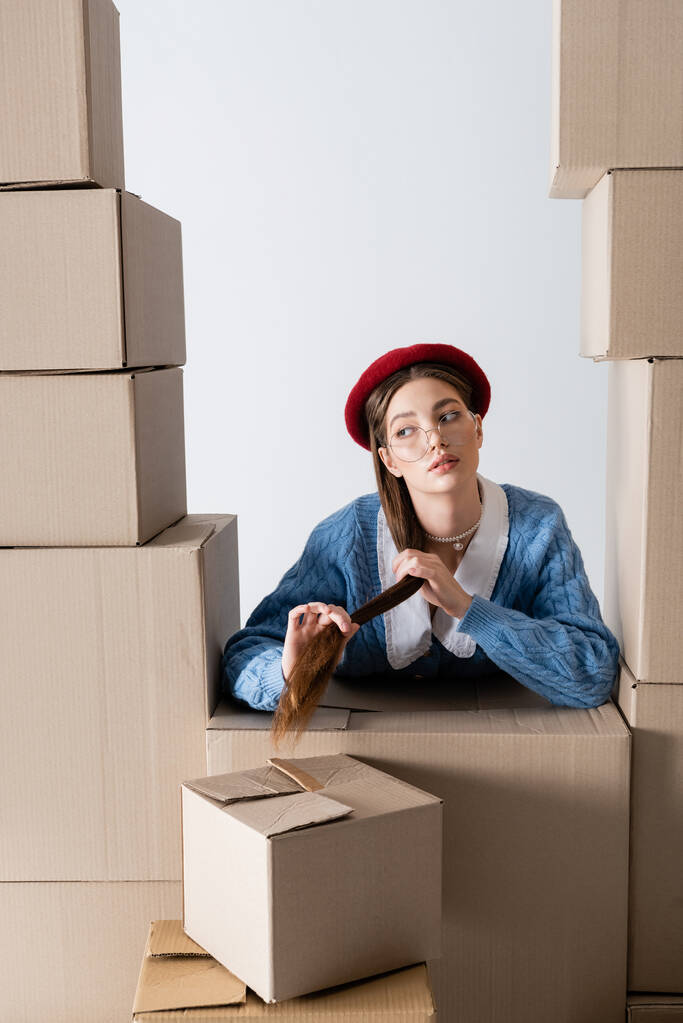 Young model in beret and eyeglasses touching hair between cardboard boxes isolated on white  - 写真・画像
