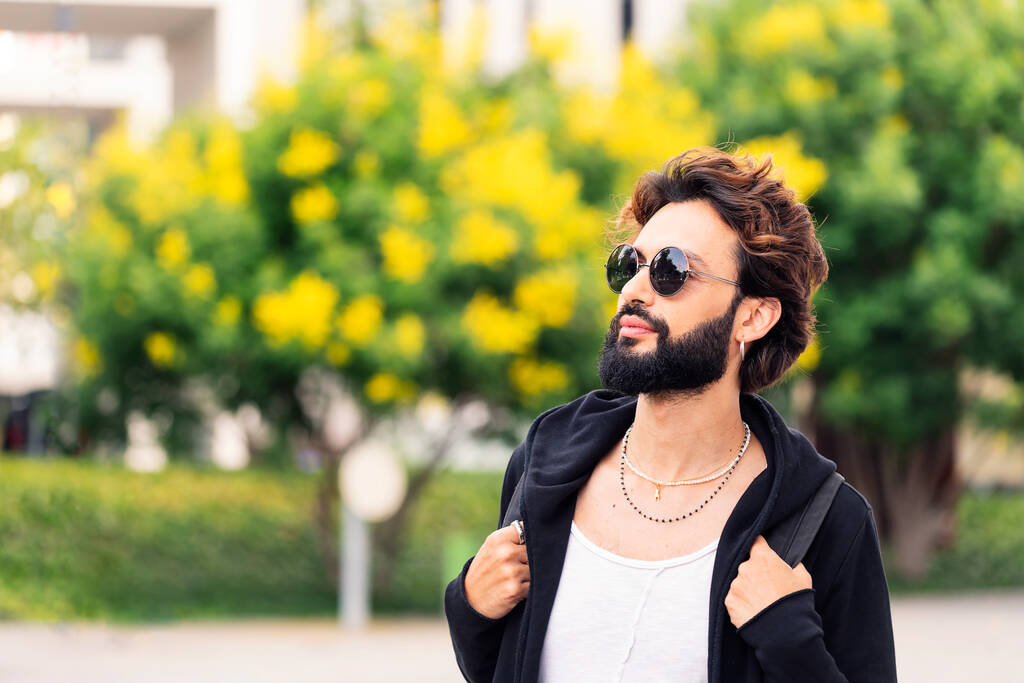 portrait of a stylish young college student with beard and sunglasses walking happy by the university campus, concept of youth and urban life style, copy space for text - 写真・画像
