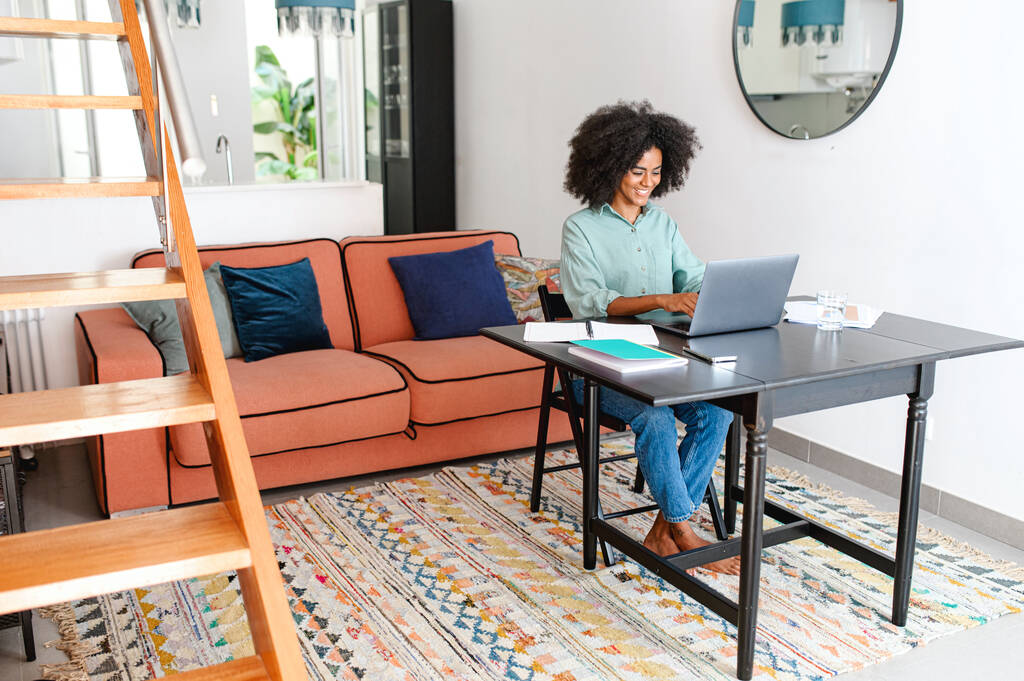 Smiling young multiracial female entrepreneur sitting at the table with laptopat home office, studying or working from home, watching an online webinar or a course, typing email - 写真・画像