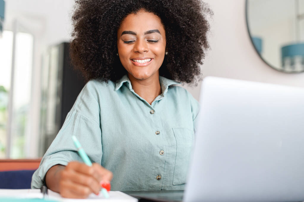 Smiling african American female student sits at desk watch webinar taking notes, multiracial young woman with curly hair working or studying using laptop computer, write in notebook - Фото, зображення