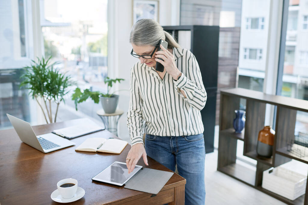 Staying connected to the best opportunities for her business. a mature businesswoman talking on a cellphone while using a digital tablet in an office - 写真・画像