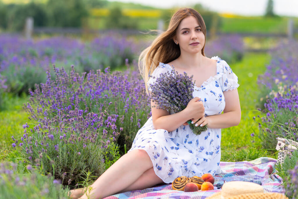 Smiling young woman posing with bunch of fresh lavender in her arms as she sits on a rug on the farm enjoying a picnic - Zdjęcie, obraz