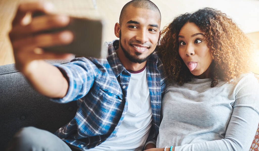Funny and silly couple taking selfie and making goofy face at phone for social media post while relaxing. Boyfriend and girlfriend having fun and enjoying time together at home and sharing it online. - Fotoğraf, Görsel