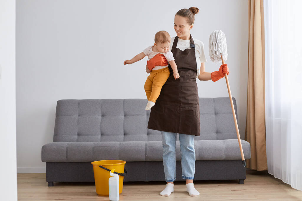 Portrait of smiling positive woman wearing brown apron with mop and little toddler daughter in hands cleaning in living room, housewife in maternity leave.. - Fotó, kép