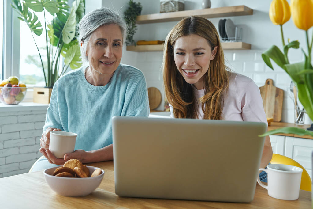Щаслива мати і її доросла дочка використовують ноутбук, проводячи час на кухні разом
 - Фото, зображення
