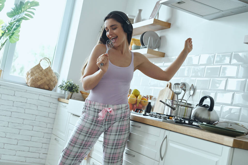 Happy young woman using whisk as microphone and dancing while cooking at the kitchen - Foto, Imagem