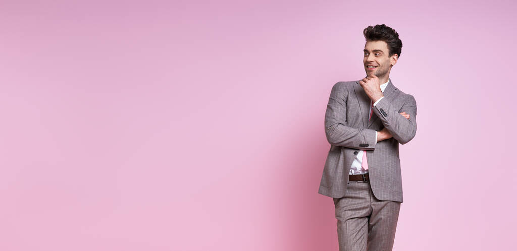 Веселый молодой человек в полном костюме, держась за подбородок, стоя на розовом фоне - Фото, изображение