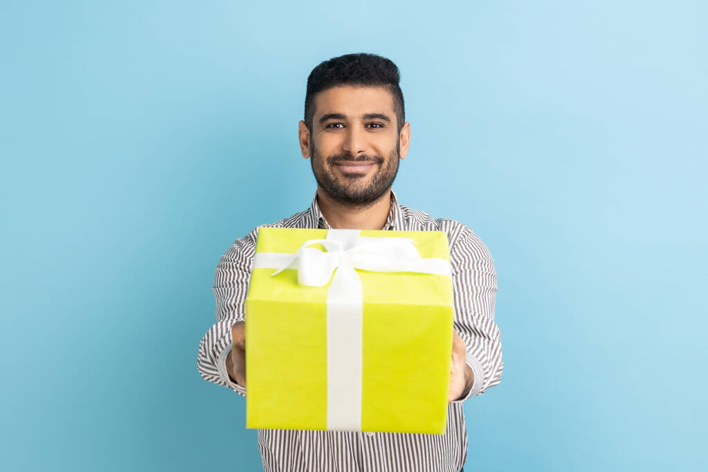 Portrait of smiling bearded businessman giving wrapped yellow gift box and smiling at camera, happy holiday, wearing striped shirt. Indoor studio shot isolated on blue background. - Foto, Imagem