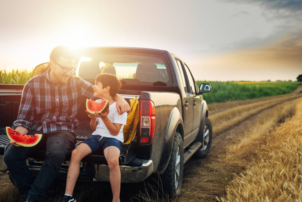 father and son sits on truck of car and eating watermelon. they are outdoor in wheat field - Photo, image