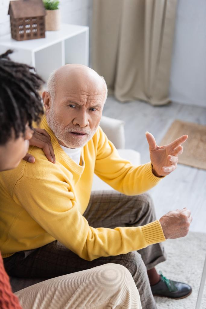 Недовольный пожилой человек, разговаривающий дома с размытым африканским внуком  - Фото, изображение