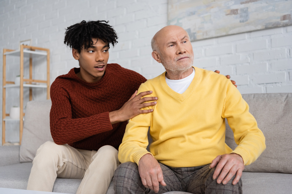 African american grandson hugging disappointed grandpa on couch at home  - 写真・画像