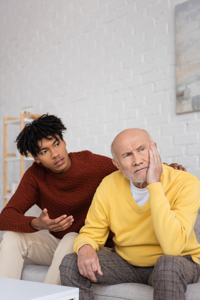 Расстроенный пожилой человек сидит рядом с африканским внуком и разговаривает дома  - Фото, изображение