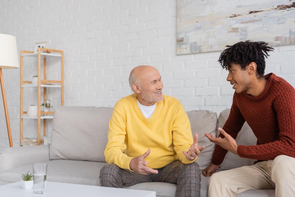 Веселый межрасовый дедушка и внук разговаривают дома на диване  - Фото, изображение